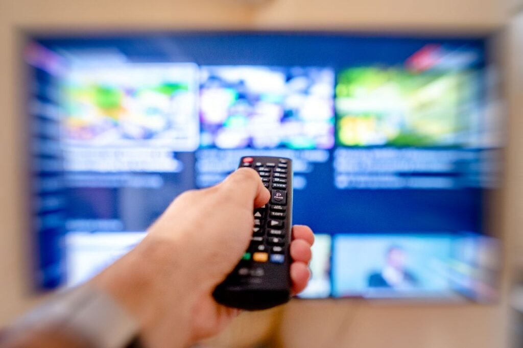 The Big3:Television Advertisements Hand holding remote control pointing at a blurred TV screen in a cozy indoor setting.